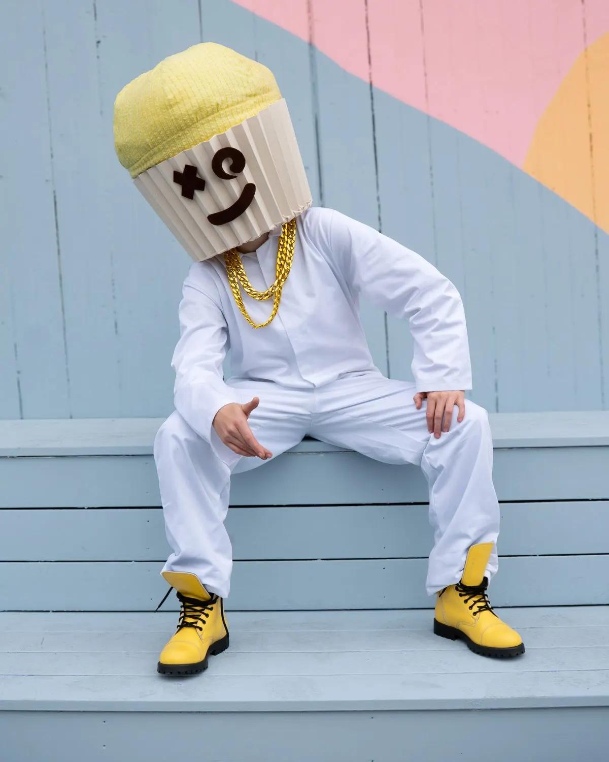 Person in a white suit and yellow boots wearing a cupcake mask, sitting on a light blue bench